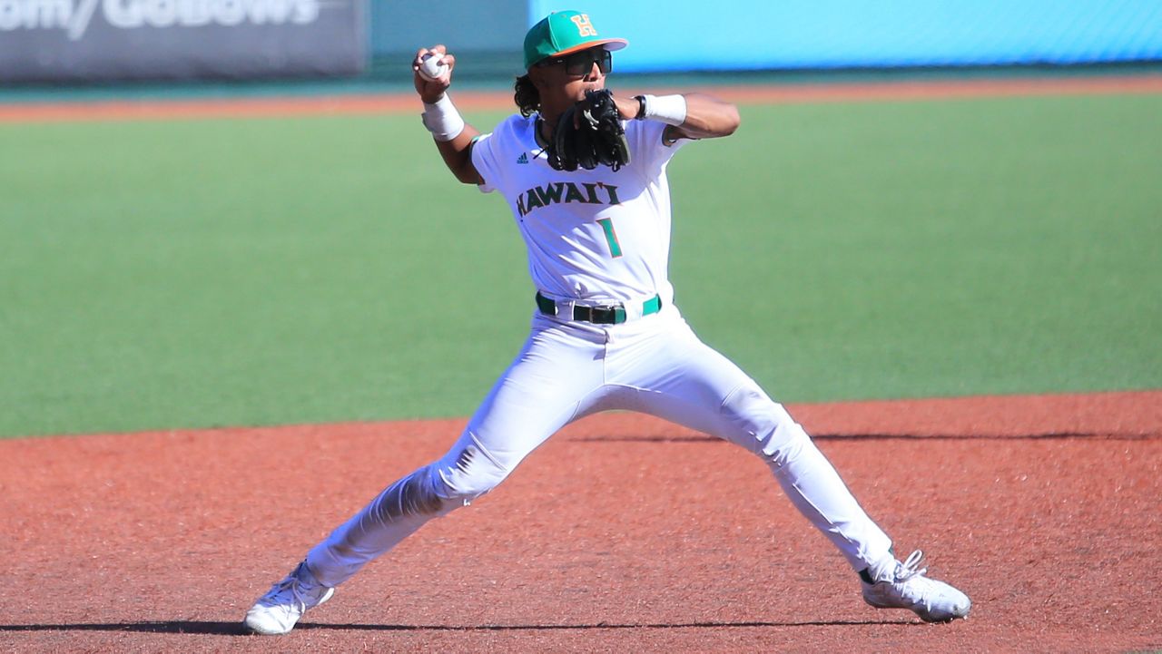 Elijah Ickes, clad in a white baseball uniform with the word "Hawaii" across the chest, cocks his arm to throw across the diamond.